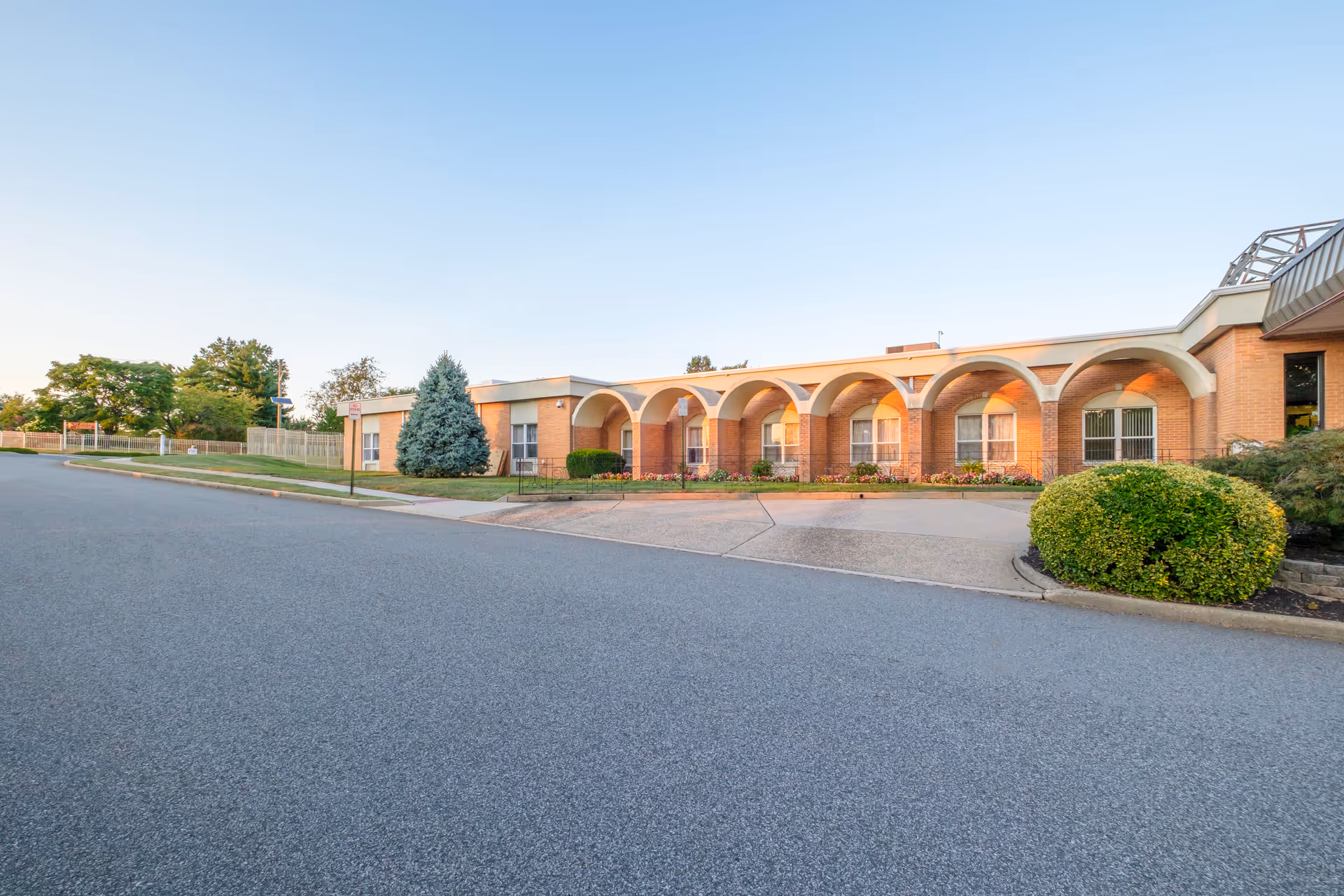 Exterior view of St Joseph's Senior Home - Assisted Living and Nursing Center showing a single-story brick building with arched architectural features, windows, and a landscaped area with bushes and a tree under a clear blue sky.