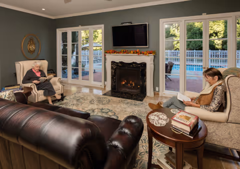 A cozy living room with two elderly women sitting in armchairs reading books. The room features a fireplace with a TV mounted above it, large windows and glass doors showing a fenced outdoor pool area, a leather sofa, and a round wooden side table with books and a doily.