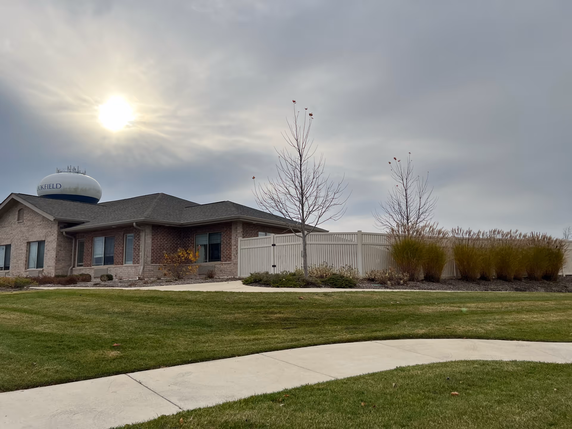 Exterior view of a single-story brick building with a fenced area and landscaped lawn under a cloudy sky with the sun partially visible. A water tower labeled 'ROCKFIELD' is seen in the background.