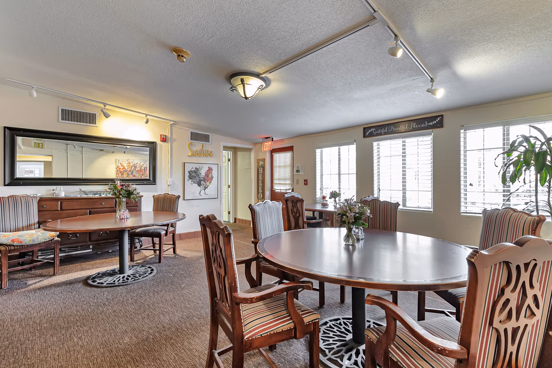 A bright dining room in a senior living facility with round wooden tables and striped cushioned chairs. There are flower vases on the tables, a large mirror on one wall, and several windows letting in natural light. Wall decorations include a framed rooster picture, a sign that says 'Sunshine', and another sign above the windows that reads 'Grateful Thankful Blessed'.
