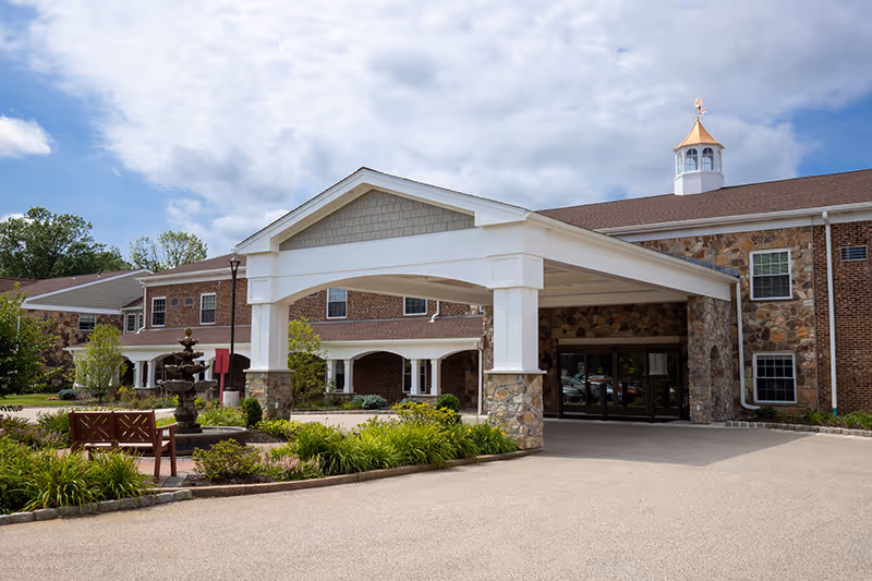 Exterior view of a senior living facility named Spring House Estates featuring a covered entrance with stone pillars, a brick and stone building facade, landscaped greenery, a fountain, and benches under a partly cloudy sky.