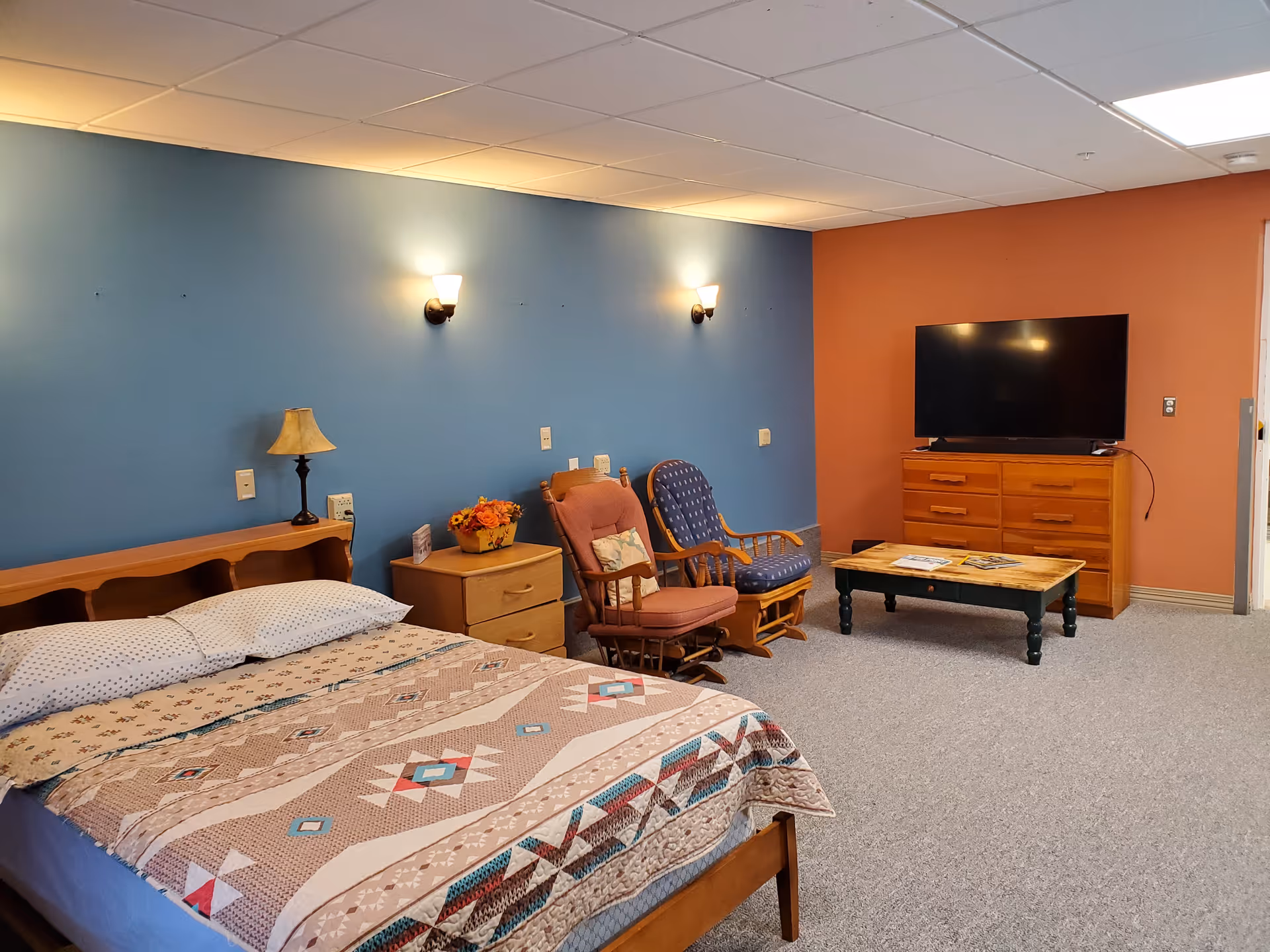 A senior living facility room with a bed covered in a patterned quilt, a wooden nightstand with a lamp and a flower arrangement, two cushioned rocking chairs, a wooden coffee table with magazines, a wooden dresser, and a flat-screen TV mounted on a peach-colored wall. The opposite wall is painted blue with two wall sconces providing light.