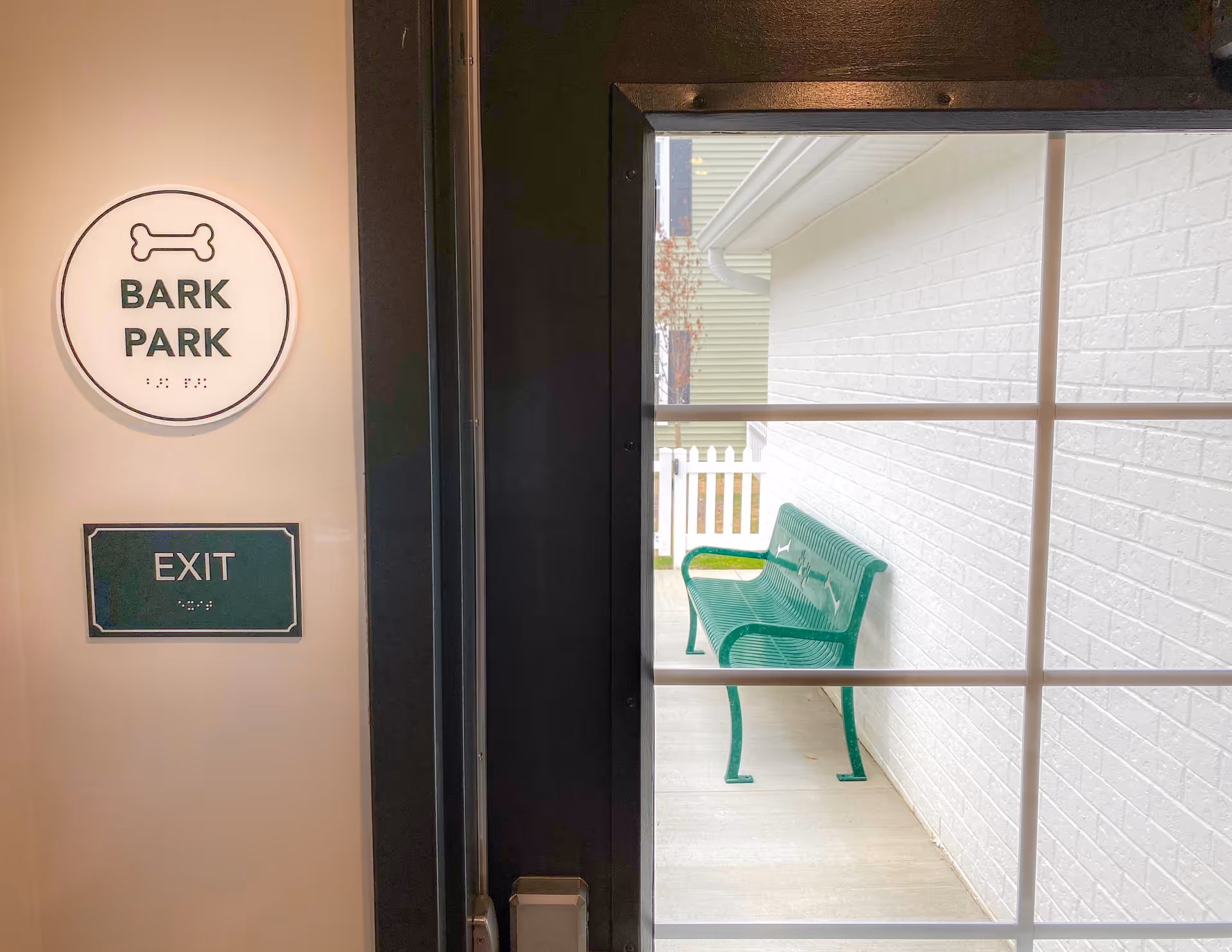 View through a glass door showing a green metal bench outside against a white brick wall. On the left side of the door, there are two signs on the wall: a round sign with a bone icon and the text 'BARK PARK', and a rectangular sign below it with the text 'EXIT'.