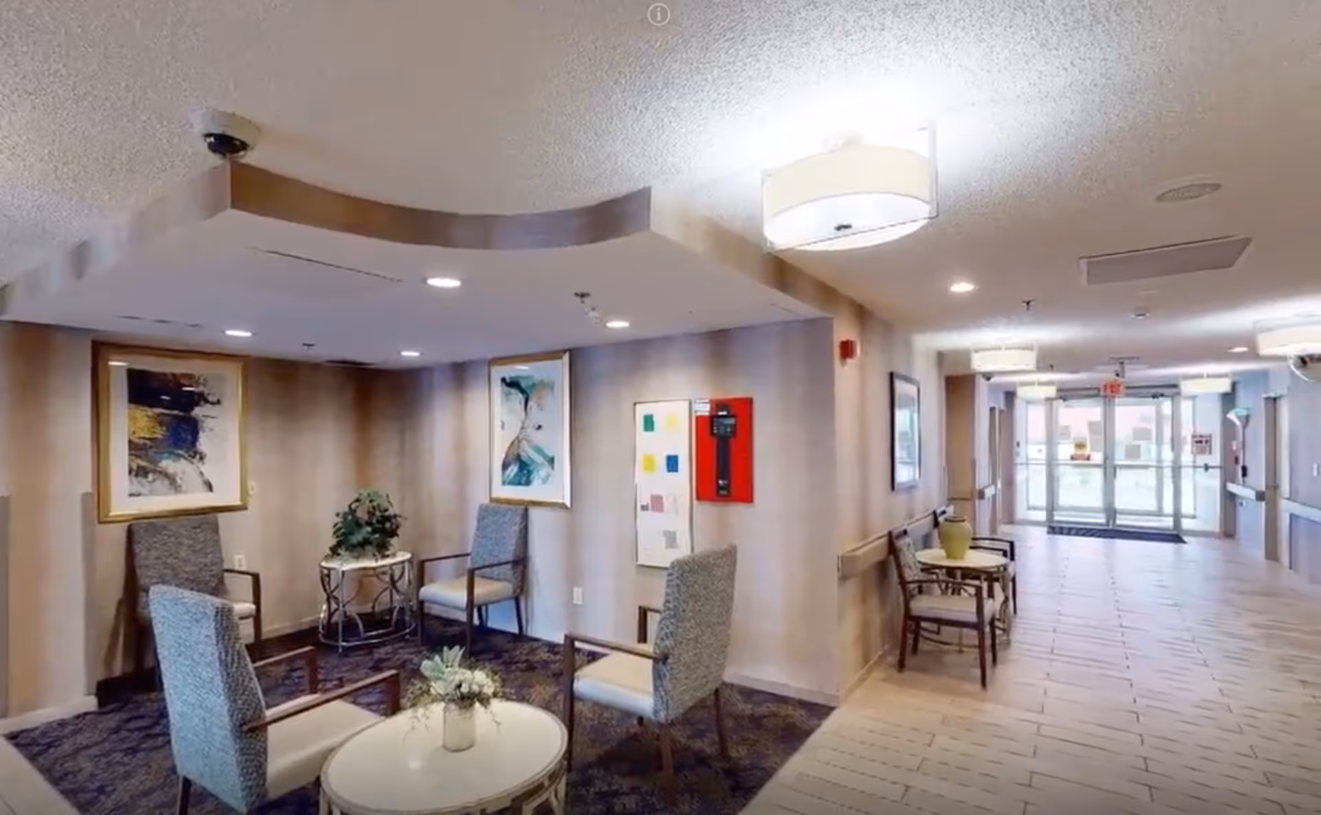 Well-lit senior living facility lobby with seating area, artwork, and a hallway leading to glass entrance doors.