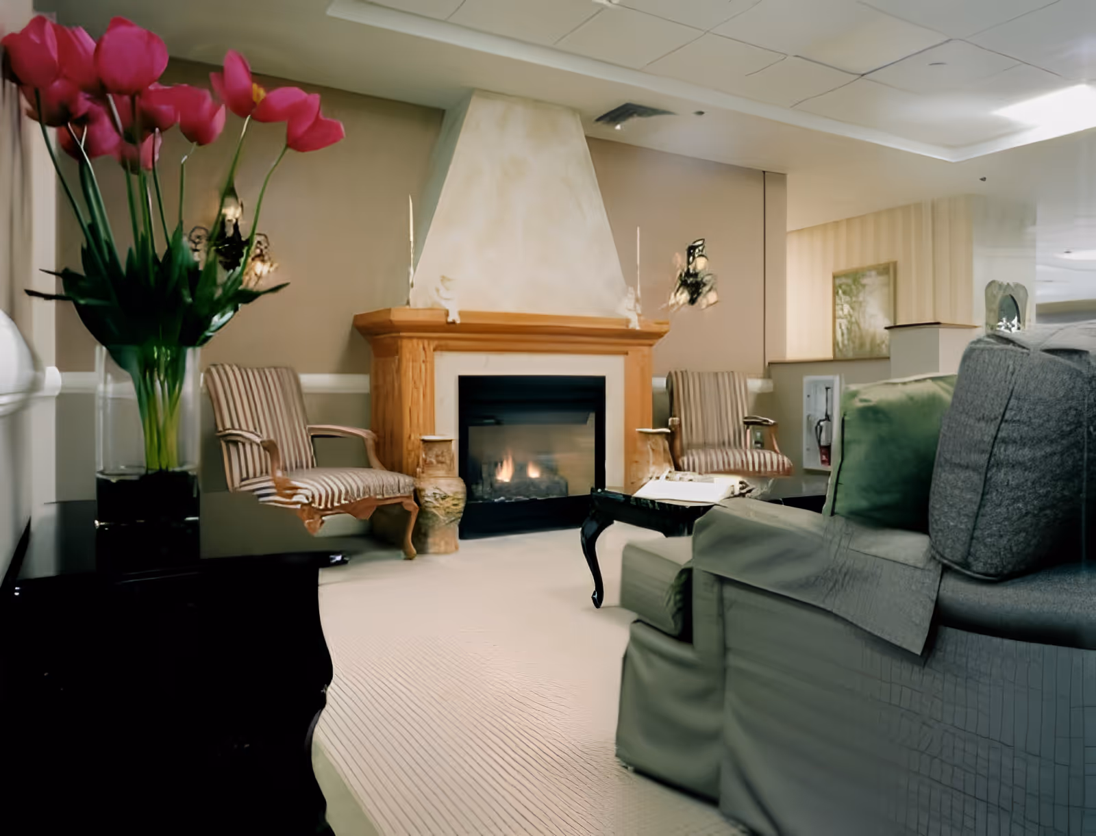 Cozy lounge area with a fireplace, upholstered chairs, a sofa, and a vase of pink tulips on a side table.