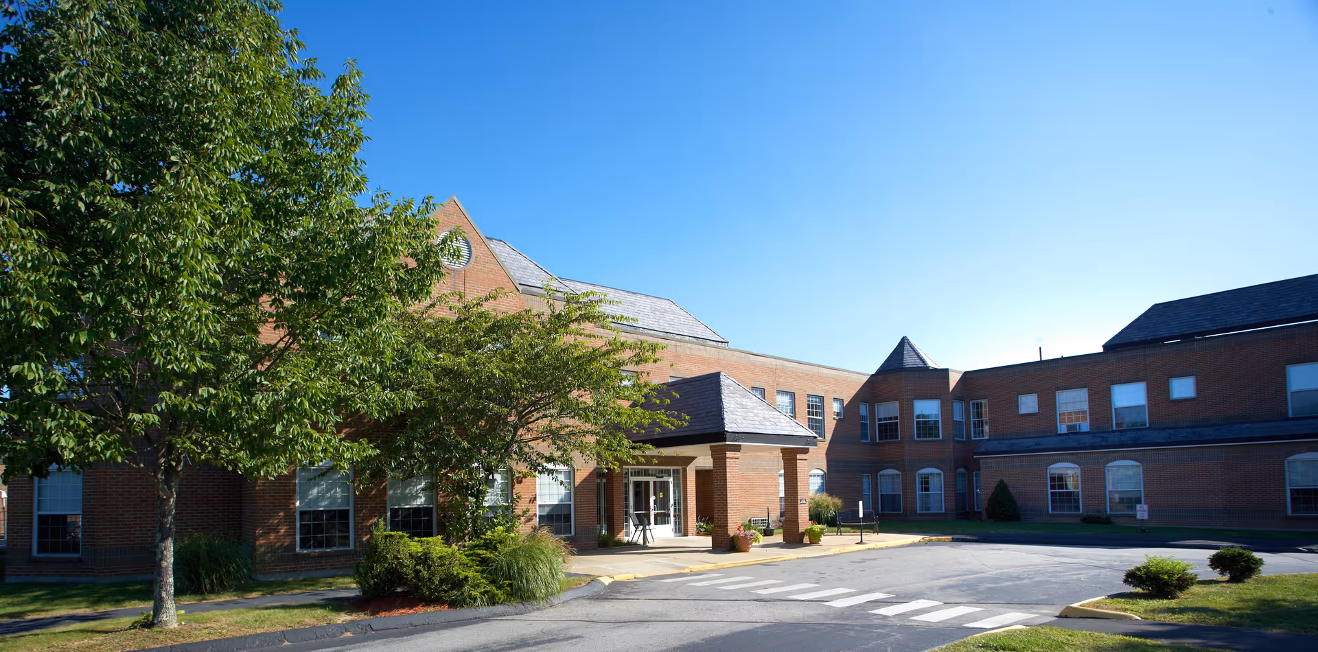 Exterior view of a two-story brick building with multiple windows, a covered entrance, and surrounding greenery including trees and shrubs under a clear blue sky.