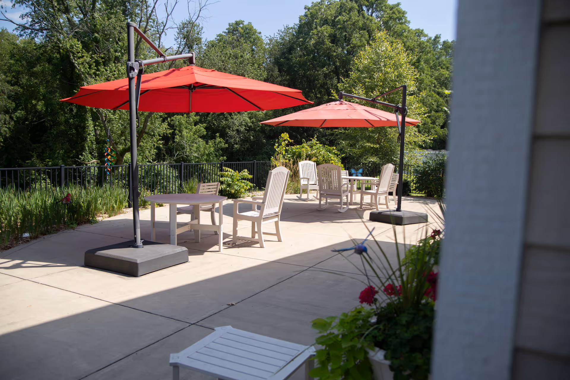 Outdoor patio area with two large red umbrellas providing shade over several white tables and chairs. The patio is surrounded by greenery and trees, with a clear blue sky overhead. There are some potted plants with flowers in the foreground.