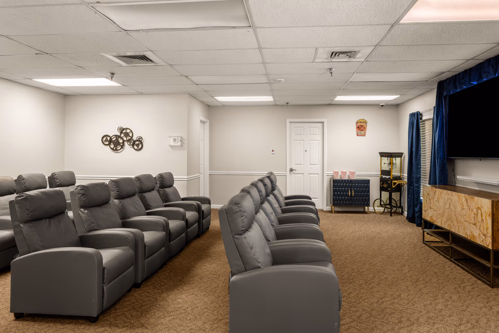 A senior living facility media room with two rows of gray reclining chairs facing a large flat-screen TV mounted on the wall. The room has beige carpet, white walls, and a popcorn machine on a small cabinet near the back door. Blue curtains frame a window to the right.