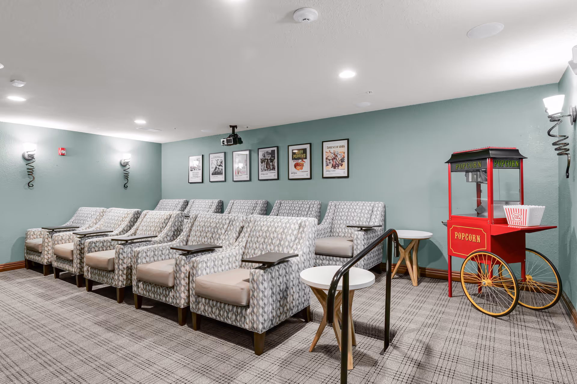 Small movie/theater room with rows of cushioned armchairs, side tables, framed posters on a green wall, and a red popcorn cart.