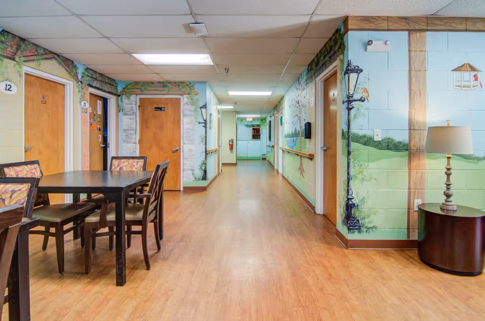 A decorated interior hallway of a senior living facility with a dining table and chairs in the foreground, mural-painted walls and room doors along the corridor.