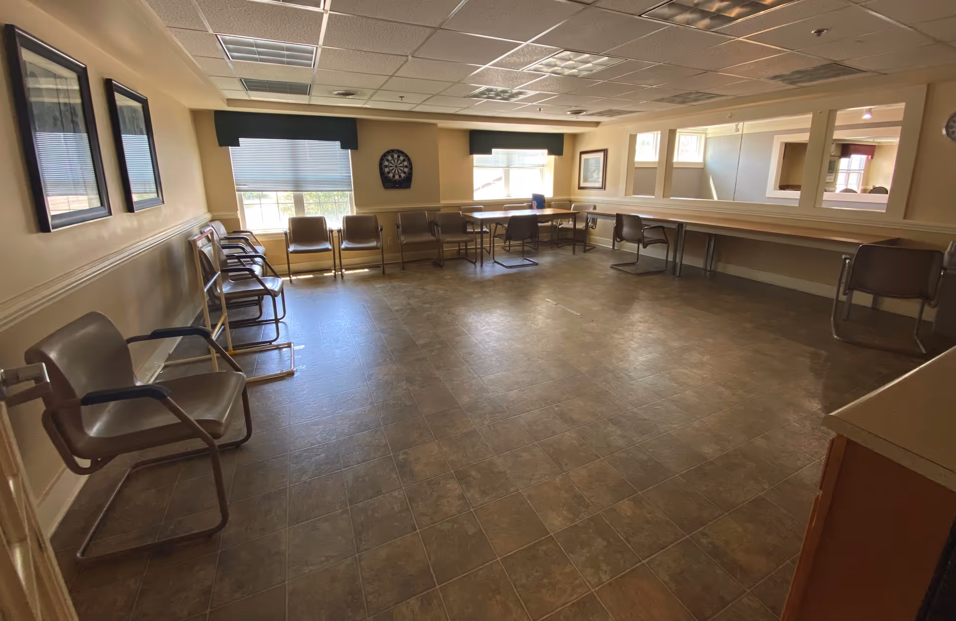 A spacious room with several brown chairs arranged along the walls and around tables. The room has large windows with blinds, a dartboard on the wall, framed pictures, and a tiled floor. The ceiling has recessed lighting and a drop ceiling design.
