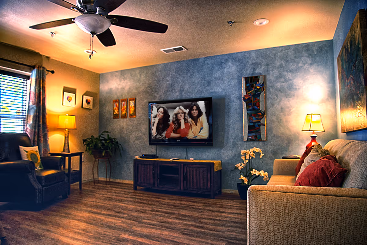 Cozy living room with a ceiling fan, a wall-mounted flat screen TV showing three women, a wooden TV stand, a beige couch with red pillows, a black armchair, two table lamps, framed artwork on the walls, a potted plant, and wood flooring.