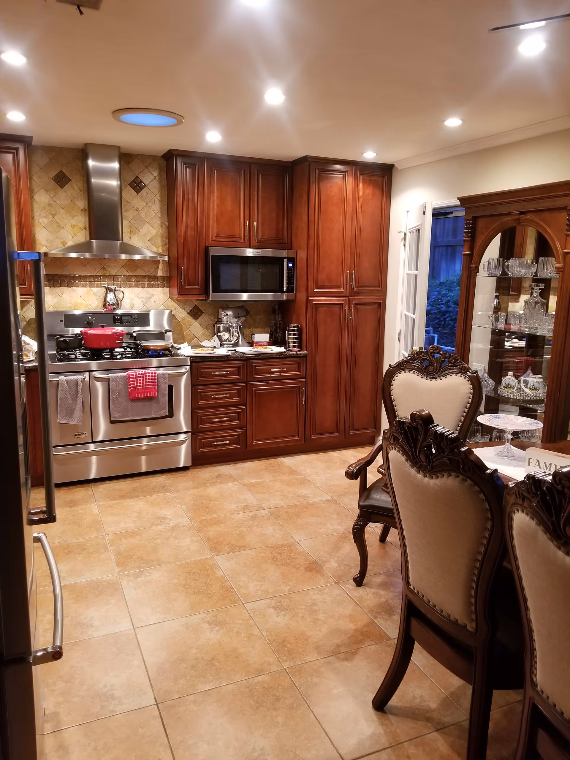 A well-lit kitchen with brown wooden cabinets, a stainless steel stove with a red pot on it, a microwave above the stove, and a tiled backsplash. To the right, there is a dining area with ornate wooden chairs upholstered in beige fabric and a glass cabinet displaying glassware. The floor is tiled with light brown tiles.