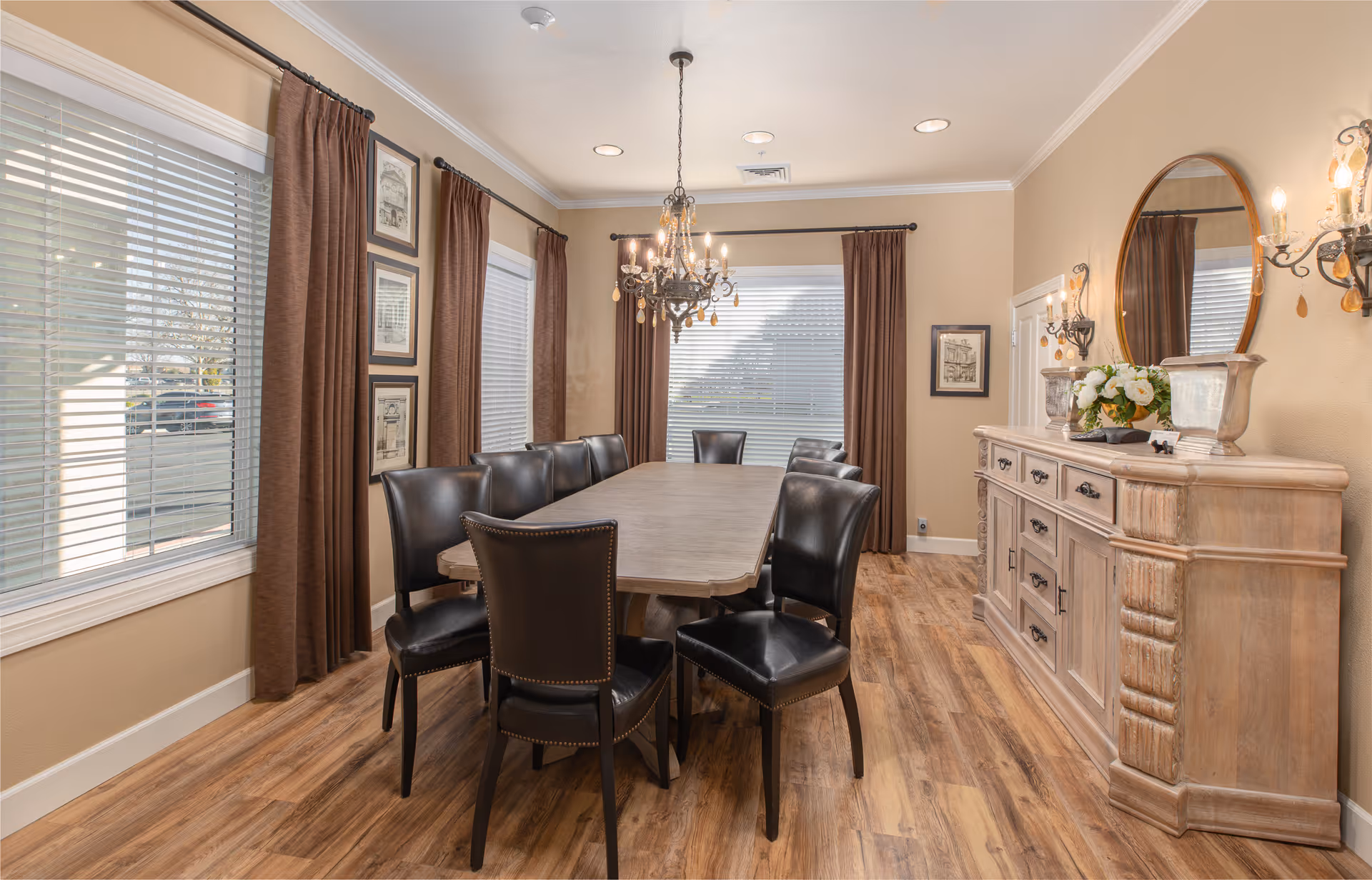 A well-lit dining room with a long wooden table surrounded by eight black leather chairs. The room features wood flooring, beige walls, three windows with brown curtains, a chandelier hanging above the table, and a wooden sideboard with decorative items including a round mirror and flower arrangement.