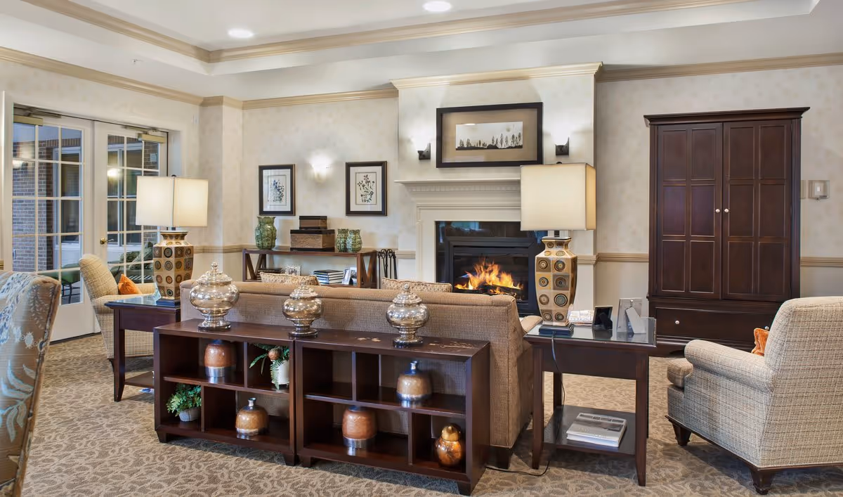 A cozy living room area in a senior living facility with a fireplace, a beige sofa, armchairs, two table lamps, decorative vases, framed artwork on the walls, and a wooden cabinet. The room has carpeted flooring and a glass door leading outside.