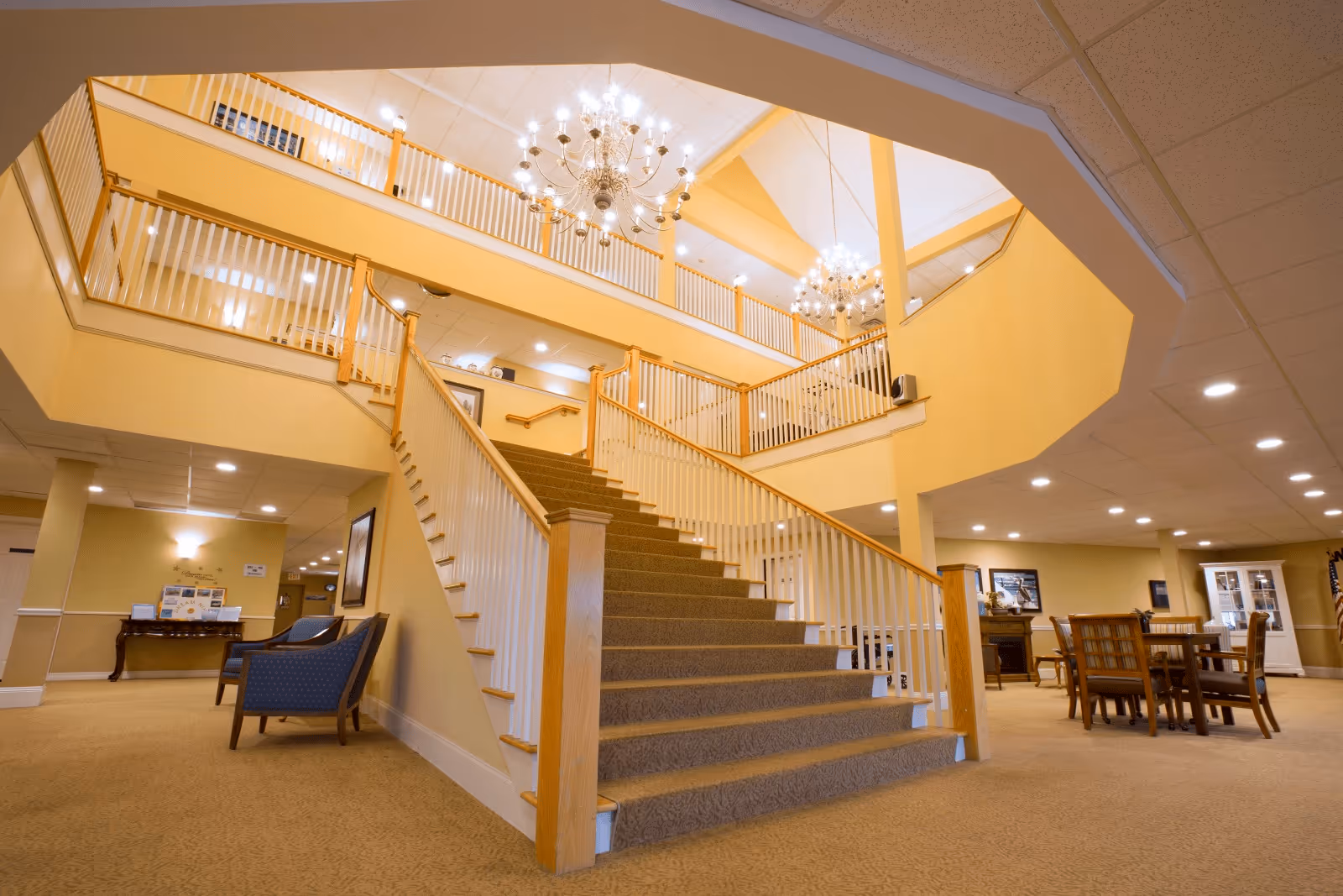 Interior view of a senior living facility with a wide carpeted staircase leading to an upper floor. The area is well-lit with chandeliers hanging from the ceiling. There are seating areas with chairs and tables on the ground floor, and the walls are painted in warm yellow tones.