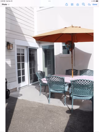 Outdoor patio area with a rectangular table covered by a pink tablecloth, surrounded by six green metal chairs. A large beige and red umbrella is positioned in the center of the table. The patio is adjacent to a white building with sliding glass doors and windows.