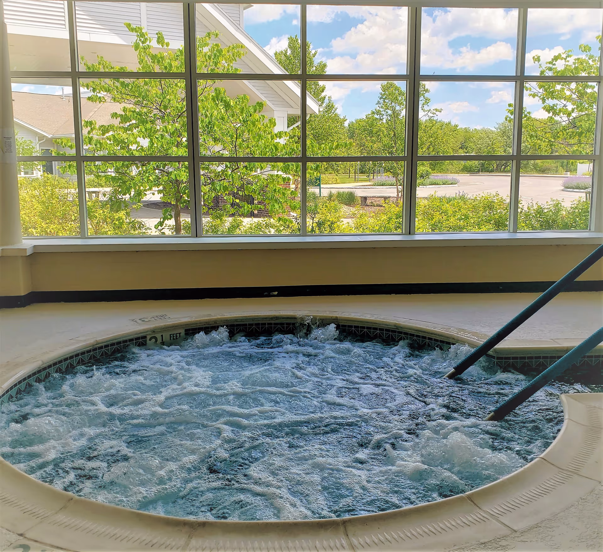 Indoor hot tub with bubbling water and metal handrails, situated next to large windows showing green trees and a partly cloudy sky outside.