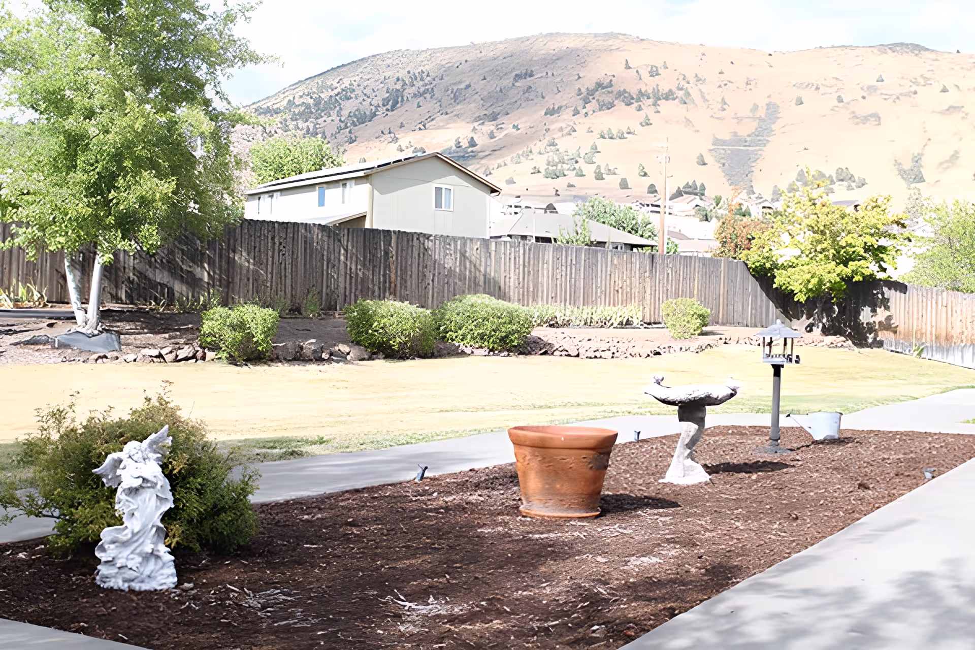 Outdoor garden area with a paved walkway, a large terracotta pot, a birdbath, a bird feeder, and a white angel statue surrounded by bushes. In the background, there is a wooden fence, trees, and a mountain under a partly cloudy sky.