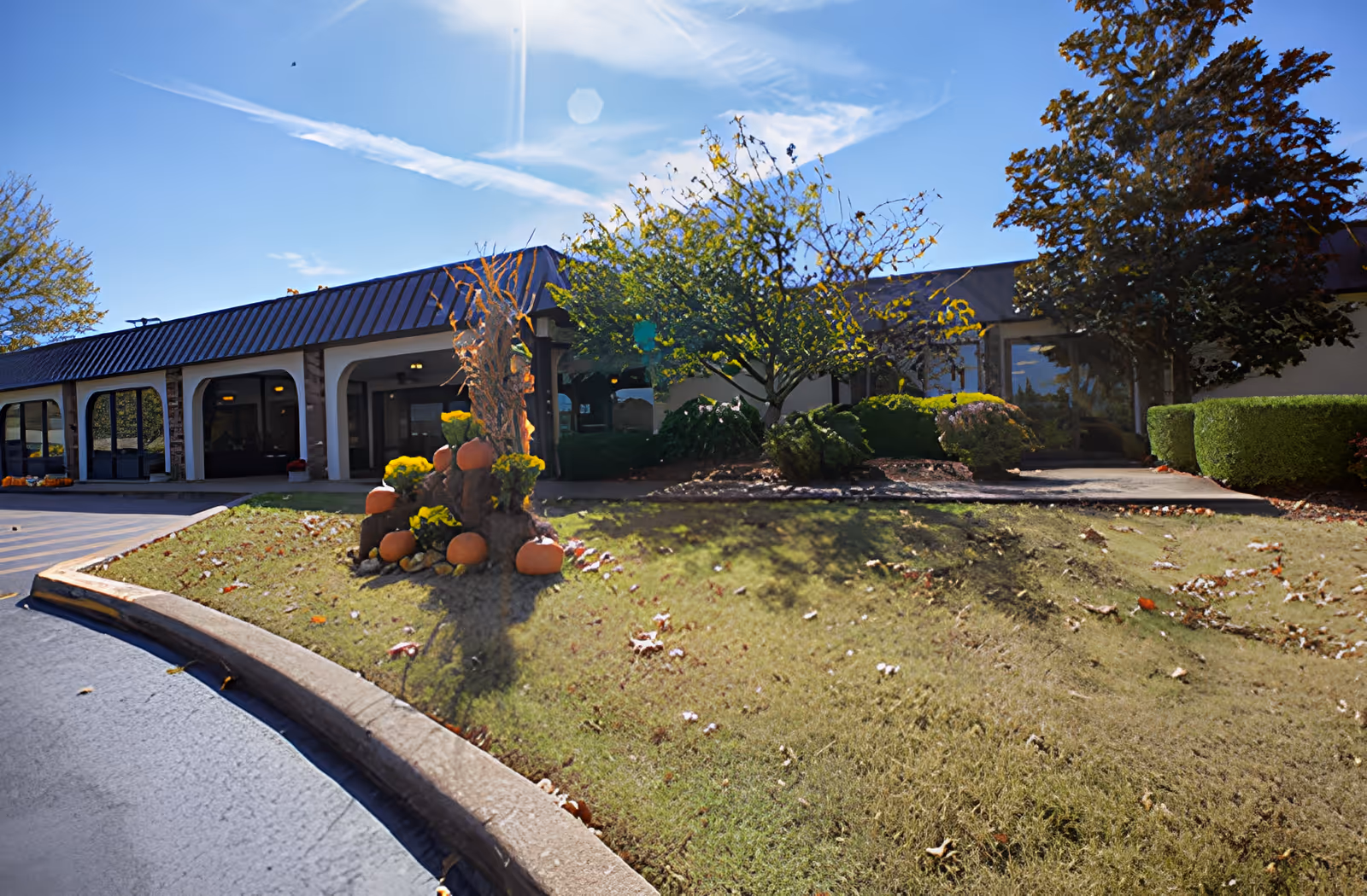 Exterior view of a single-story building with a dark roof and large windows, surrounded by landscaped bushes and trees. There is a decorative fall display with pumpkins and yellow flowers on a grassy area near the entrance. The sky is clear and sunny.