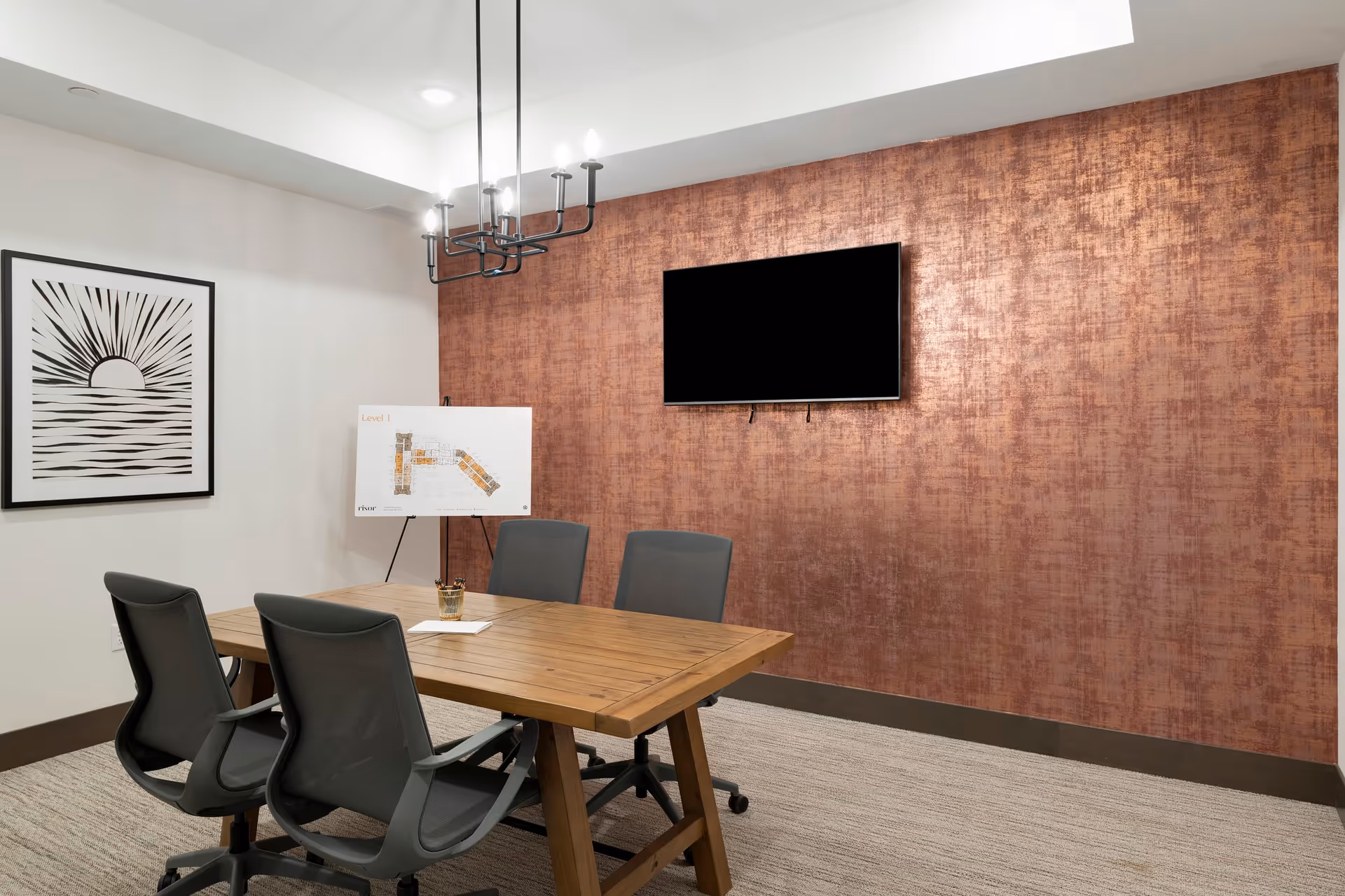 A modern meeting room with a wooden table surrounded by four gray office chairs on wheels. A flat-screen TV is mounted on a textured copper-colored wall. A framed abstract artwork depicting a sun and waves hangs on a white wall. An easel holds a floor plan labeled 'Level 1'. The room is carpeted and lit by a black chandelier with exposed bulbs.