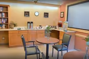 Small indoor café area with round tables and chairs, a counter holding coffee urns and a water dispenser, shelving, and framed art.