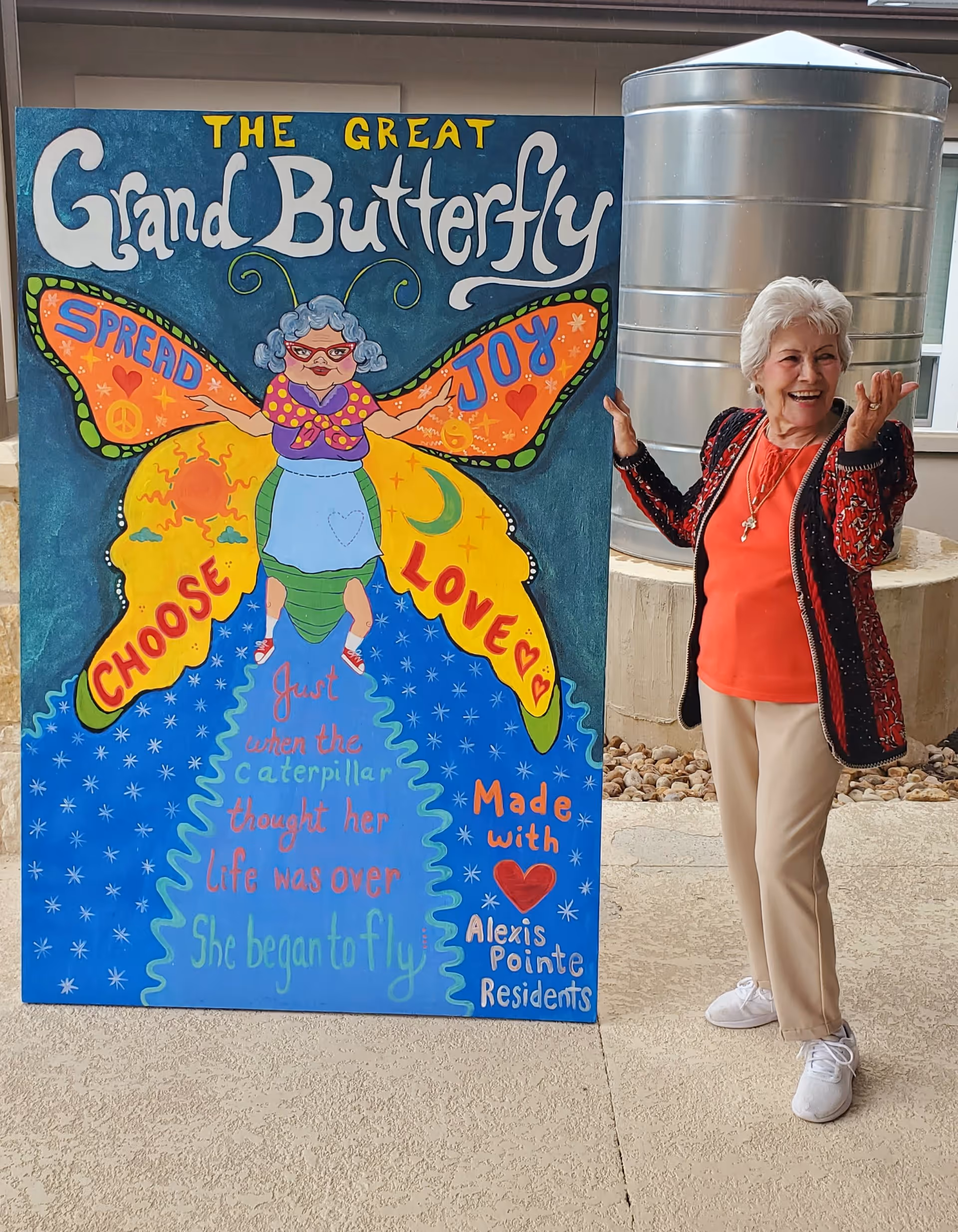 An elderly woman with white hair, wearing a red shirt, beige pants, white shoes, and a black and red jacket, stands smiling and gesturing next to a colorful painted sign. The sign features a cartoon butterfly with an elderly woman as the body and the words 'The Great Grand Butterfly' at the top. The butterfly wings have the words 'Spread Joy' and 'Choose Love' written on them. Below, the sign reads, 'Just when the caterpillar thought her life was over, she began to fly' and 'Made with love Alexis Pointe Residents.' The setting appears to be an outdoor patio area with a large metal cylindrical structure in the background.