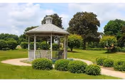 Wooden gazebo in a landscaped grassy garden with shrubs, a winding path, and trees under a partly cloudy sky.