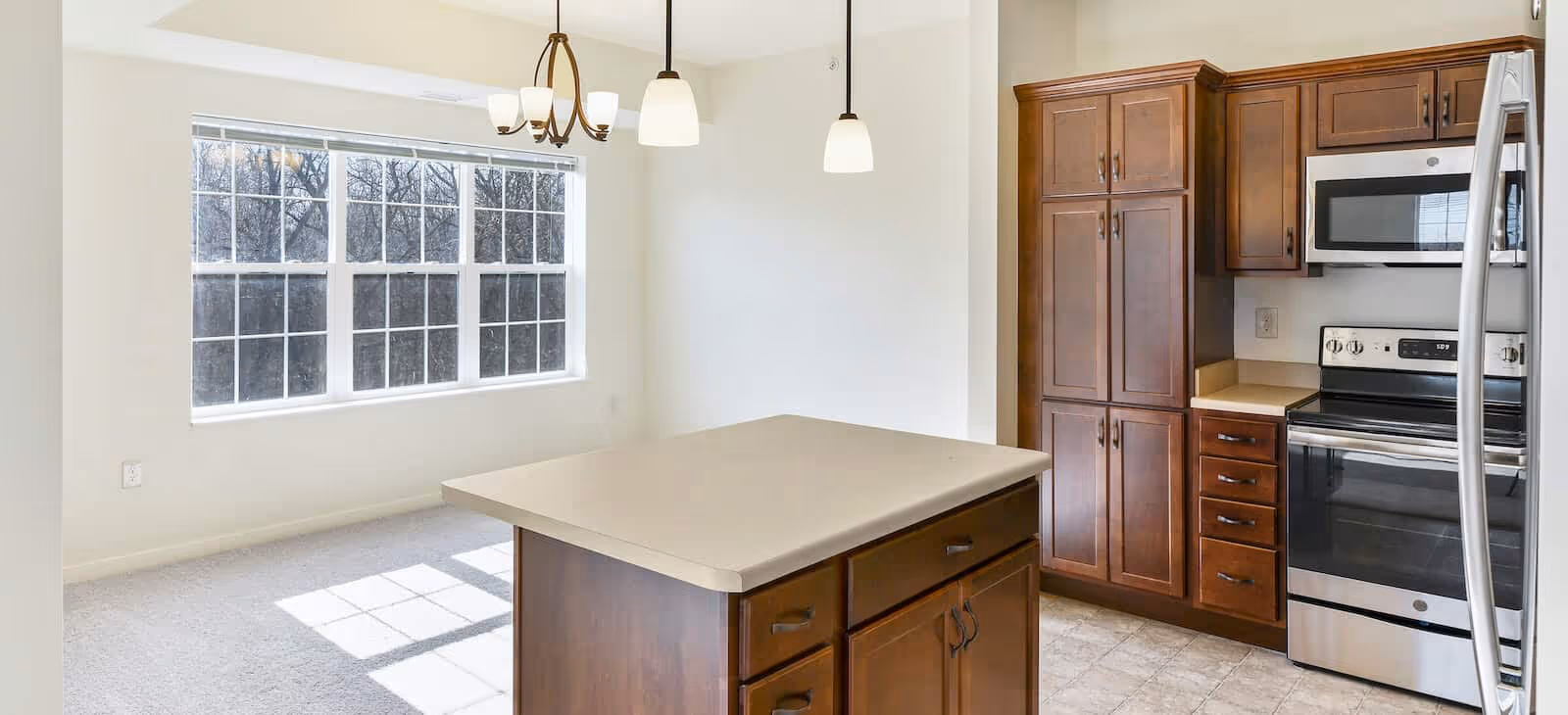 A bright kitchen area with wooden cabinets, a stainless steel stove and microwave, and a central island with drawers. Adjacent to the kitchen is a dining area with a large window letting in natural light and hanging light fixtures above.