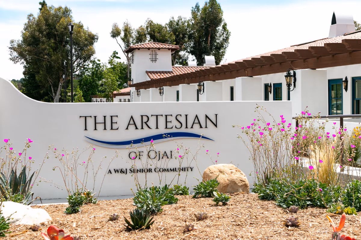 Entrance sign reading "The Artesian of Ojai" in front of white Mediterranean-style senior living buildings and landscaped grounds.