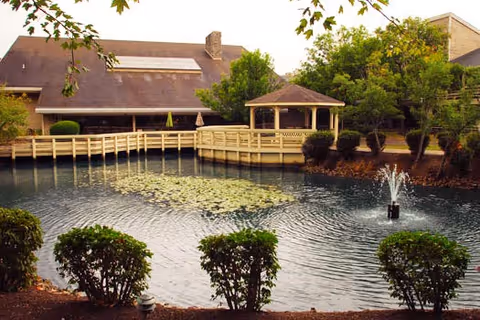 A serene outdoor scene at The Lodge Retirement Community featuring a pond with a small fountain in the center, surrounded by neatly trimmed bushes and trees. A wooden boardwalk with railings extends over the pond, leading to a gazebo. In the background, there is a building with a sloped roof and large windows.