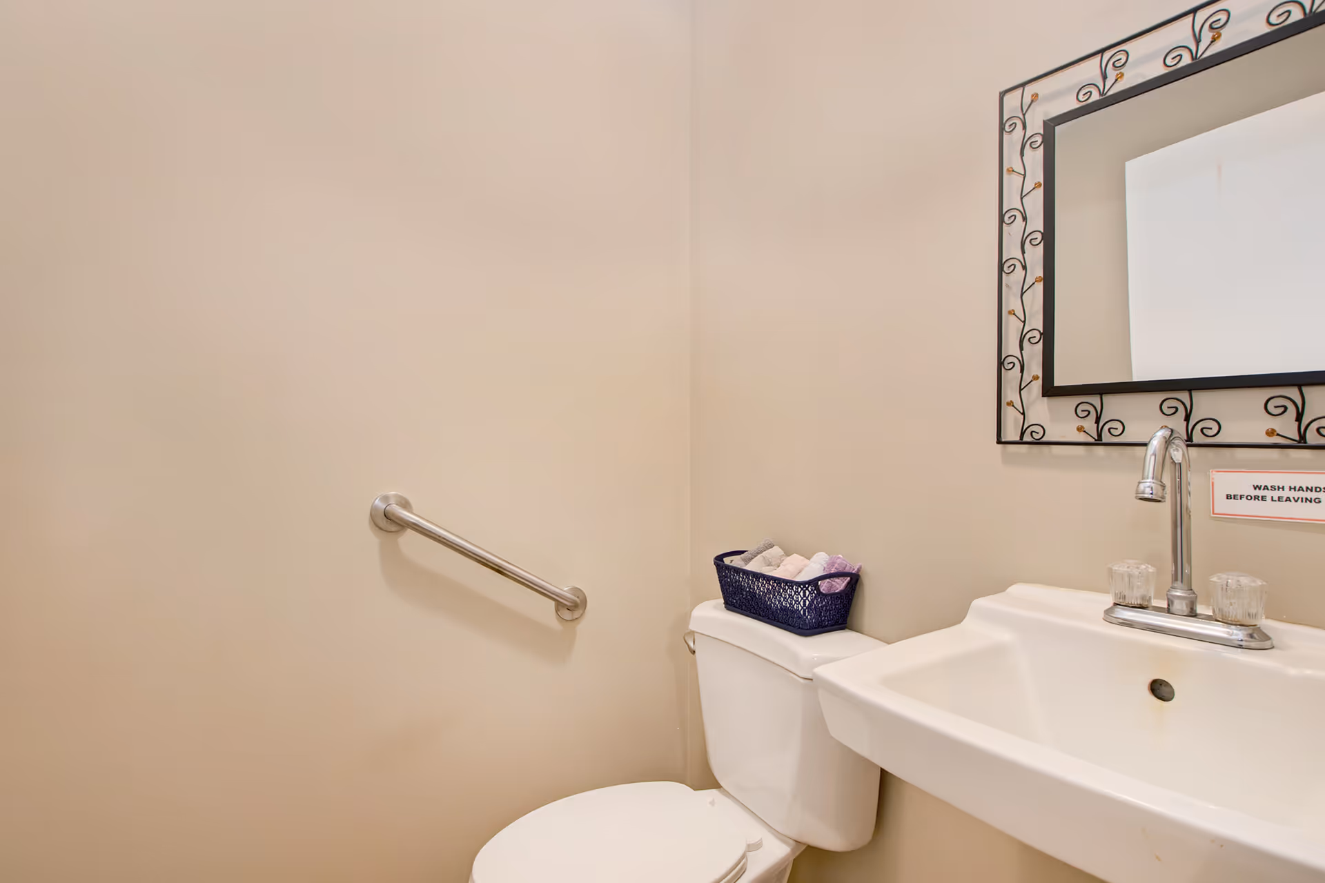 A small bathroom with a white toilet and a white sink. There is a metal grab bar on the wall next to the toilet. On top of the toilet tank is a basket with rolled towels. Above the sink is a decorative framed mirror and a sign that reads 'WASH HANDS BEFORE LEAVING'.