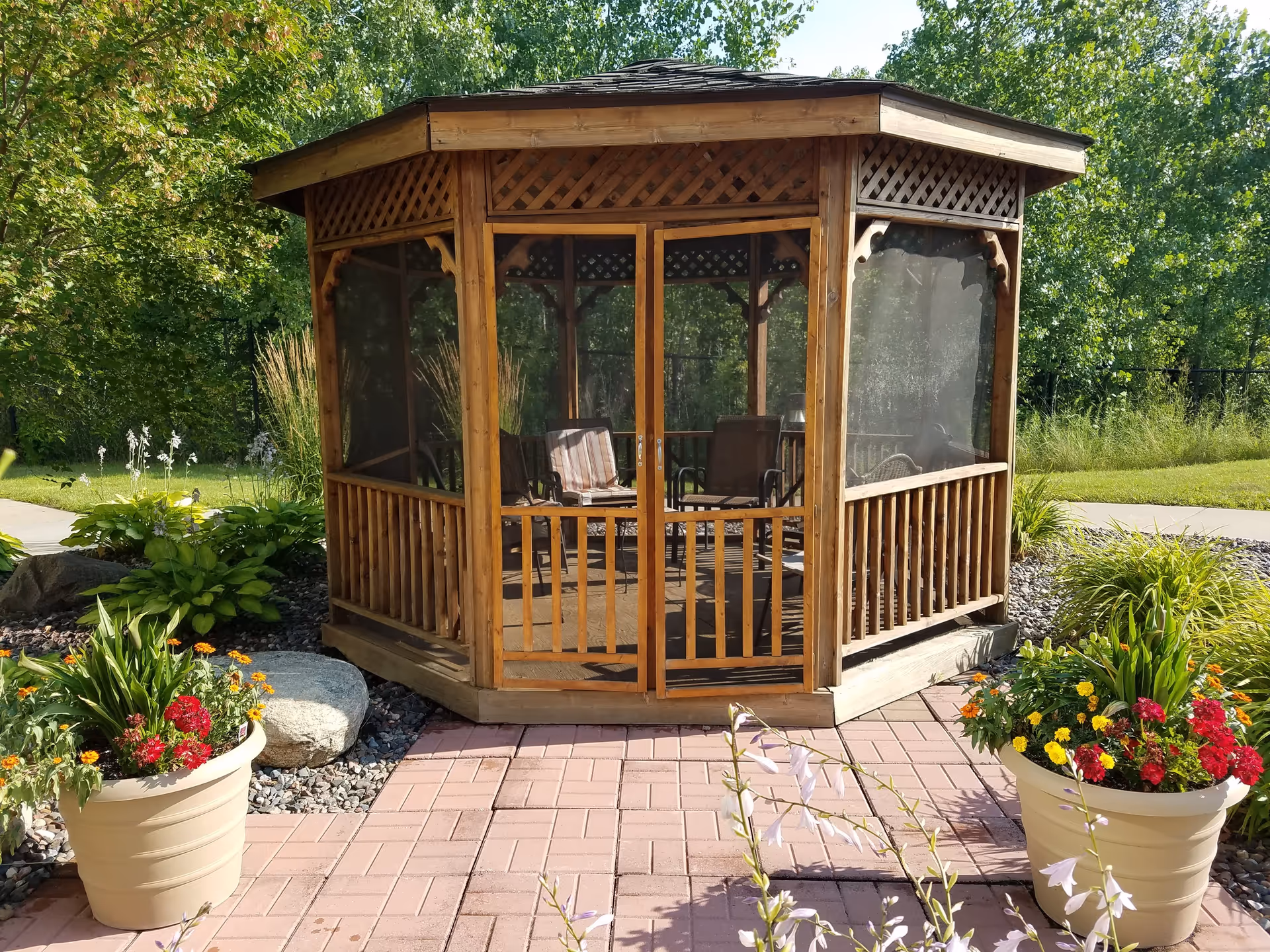 Wooden screened gazebo on a brick patio surrounded by potted flowers and landscaped greenery.