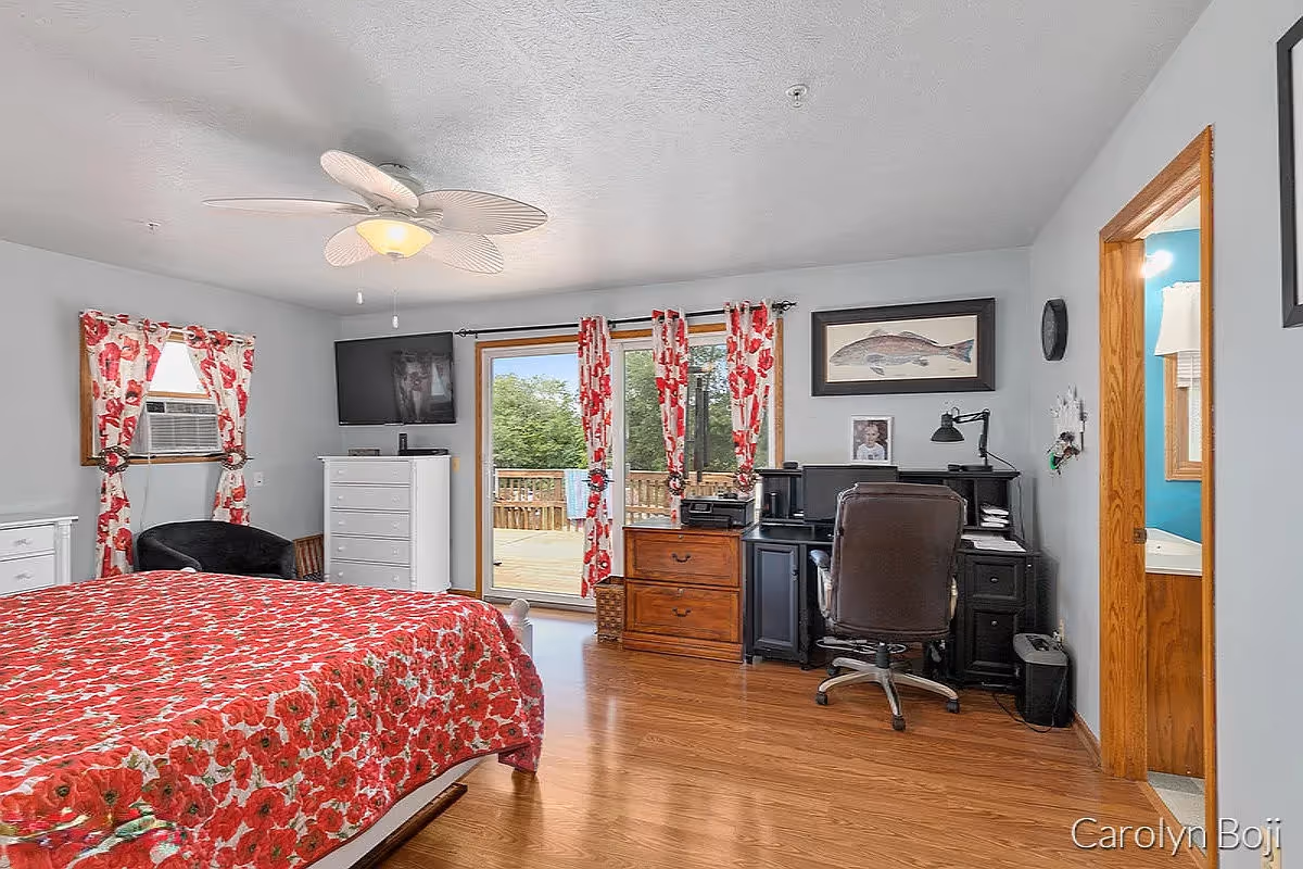 A bedroom with a bed covered in a red floral quilt, a ceiling fan with leaf-shaped blades, a window with red floral curtains, a white dresser with a TV mounted above it, a black chair, a wooden chest of drawers, and a black desk with a computer and office chair. A sliding glass door leads to an outdoor deck with greenery visible outside. There is an open doorway to a bathroom with blue walls and a white sink.