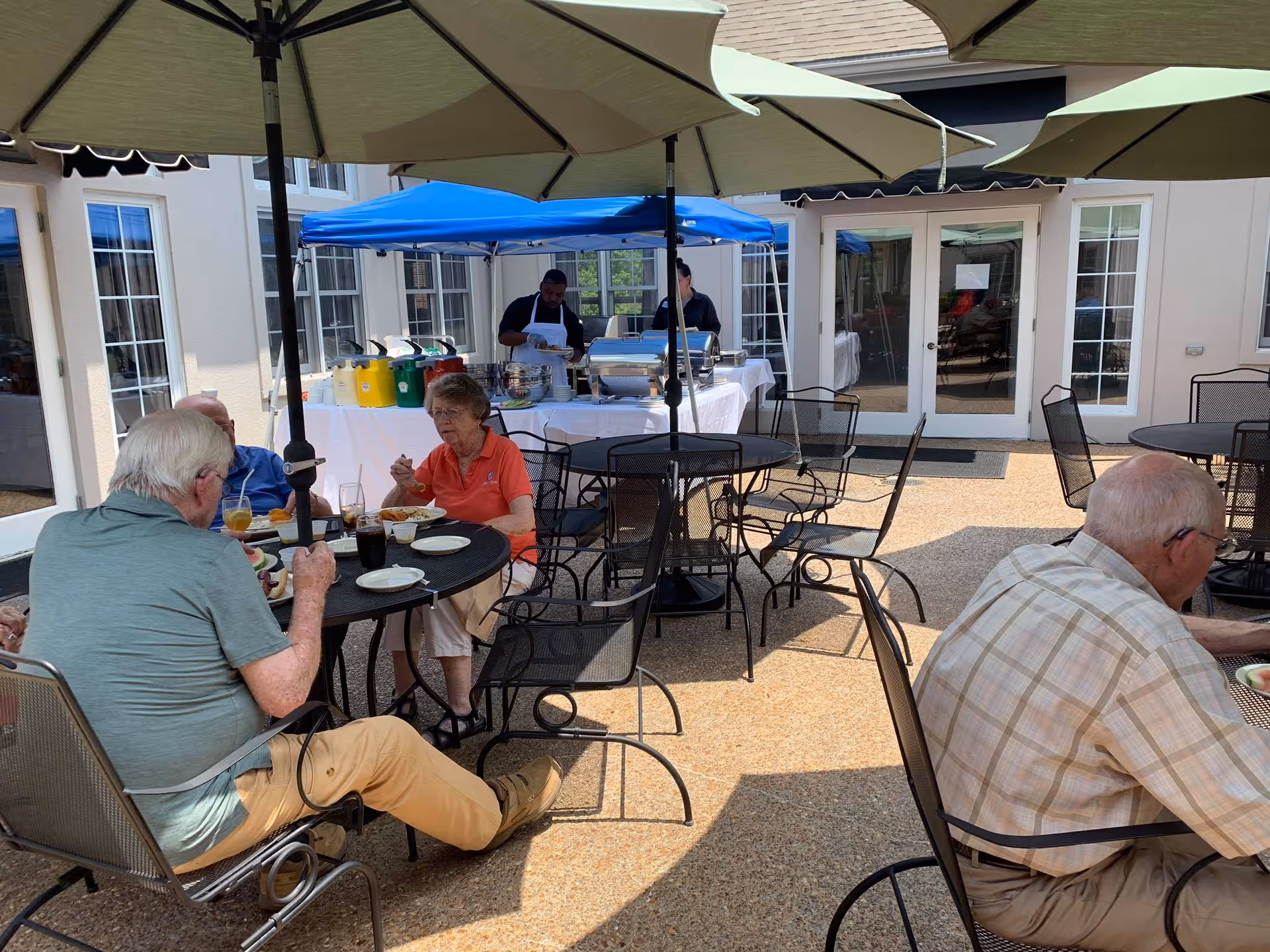 Several elderly people sitting at outdoor metal tables with umbrellas, eating and drinking. In the background, two staff members stand behind a table with food and drink dispensers under a blue canopy. The setting appears to be a patio area of a senior living facility.