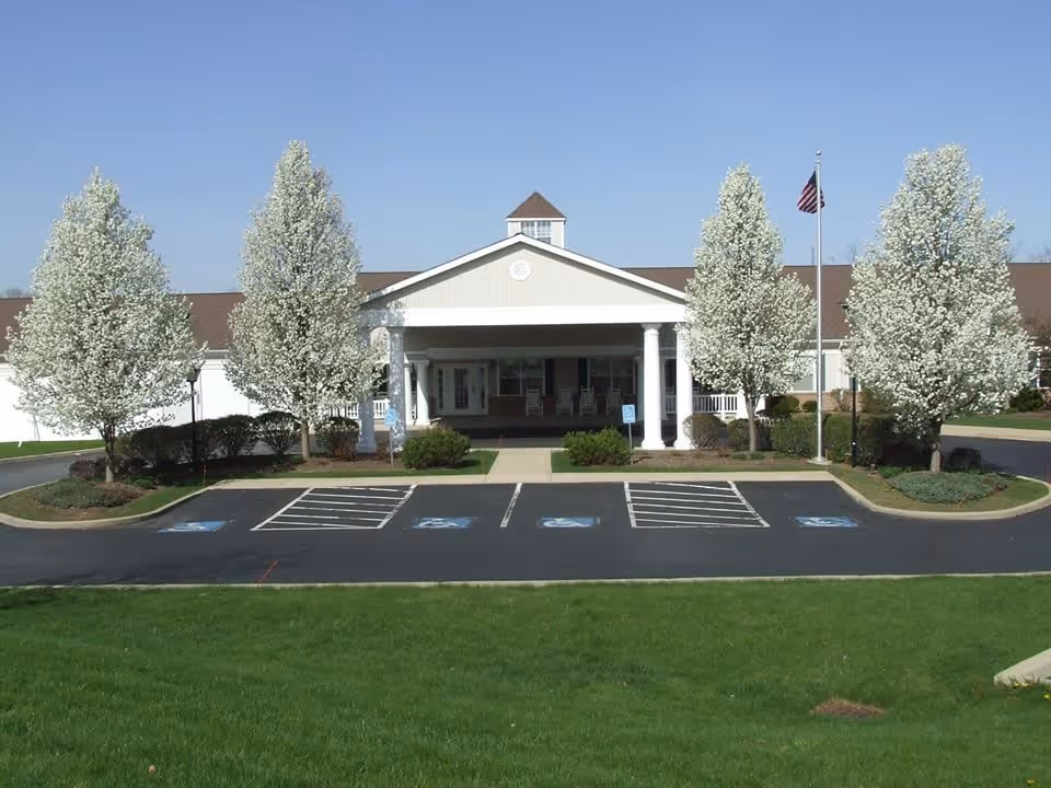 Front exterior view of a single-story building with a covered entrance supported by white columns. There are five flowering trees with white blossoms lining the driveway, which has several handicap parking spaces. An American flag is flying on a flagpole near the entrance.