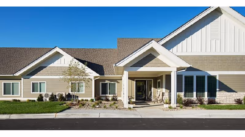 Single-story beige senior living building with a covered entryway, columns, and windows under a clear blue sky.