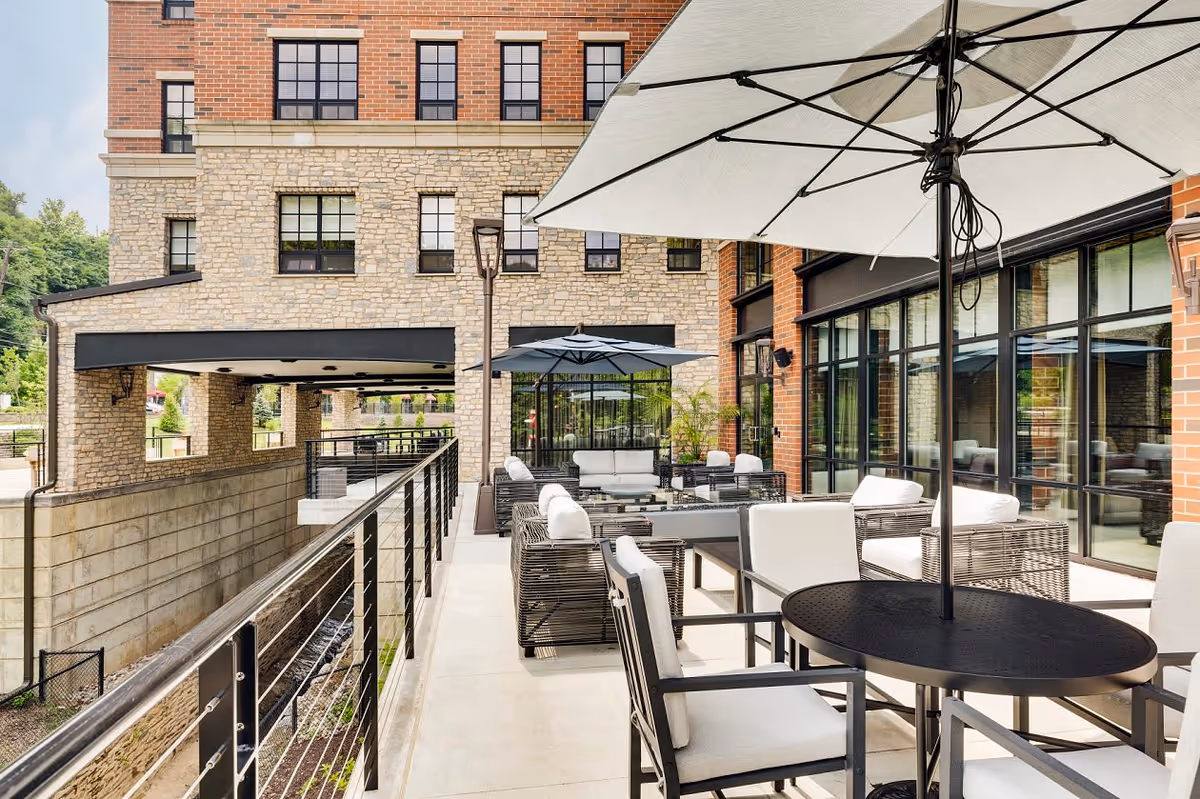 Outdoor patio area at The Residence at Bala Cynwyd featuring modern seating with cushioned chairs and sofas, black metal tables, large umbrellas for shade, and a railing overlooking a lower level. The building exterior is made of brick and stone with large windows.