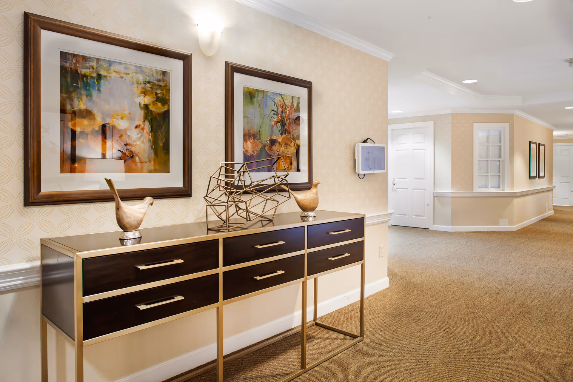 A well-lit hallway in a senior living facility with beige patterned wallpaper and carpeted floor. A modern console table with six drawers and metallic frame is against the wall, decorated with two bird figurines and a geometric metal sculpture. Above the table are two framed abstract paintings. The hallway extends with white doors and additional framed artwork visible in the distance.