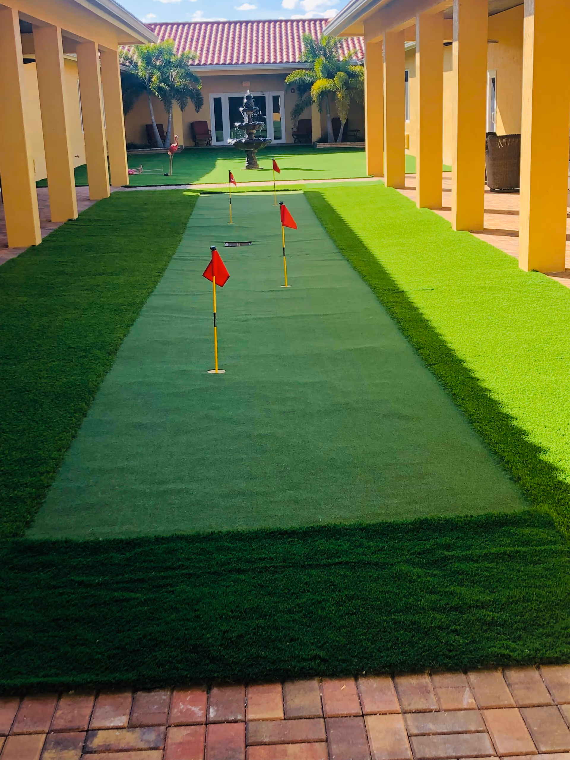 Outdoor courtyard area with a putting green featuring four small red flags. The courtyard is surrounded by yellow building walls with columns and palm trees, and there is a multi-tiered water fountain in the background.
