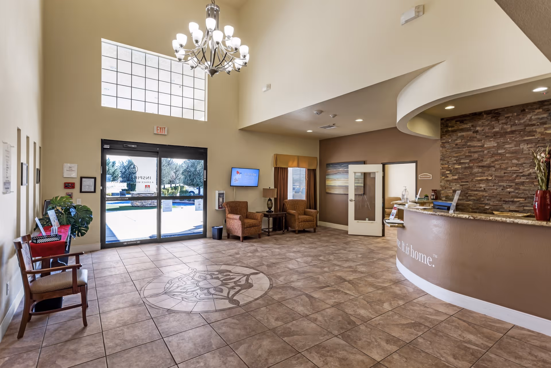 Spacious senior living lobby with tiled floor, curved reception desk, seating area and glass entrance doors under a large chandelier.
