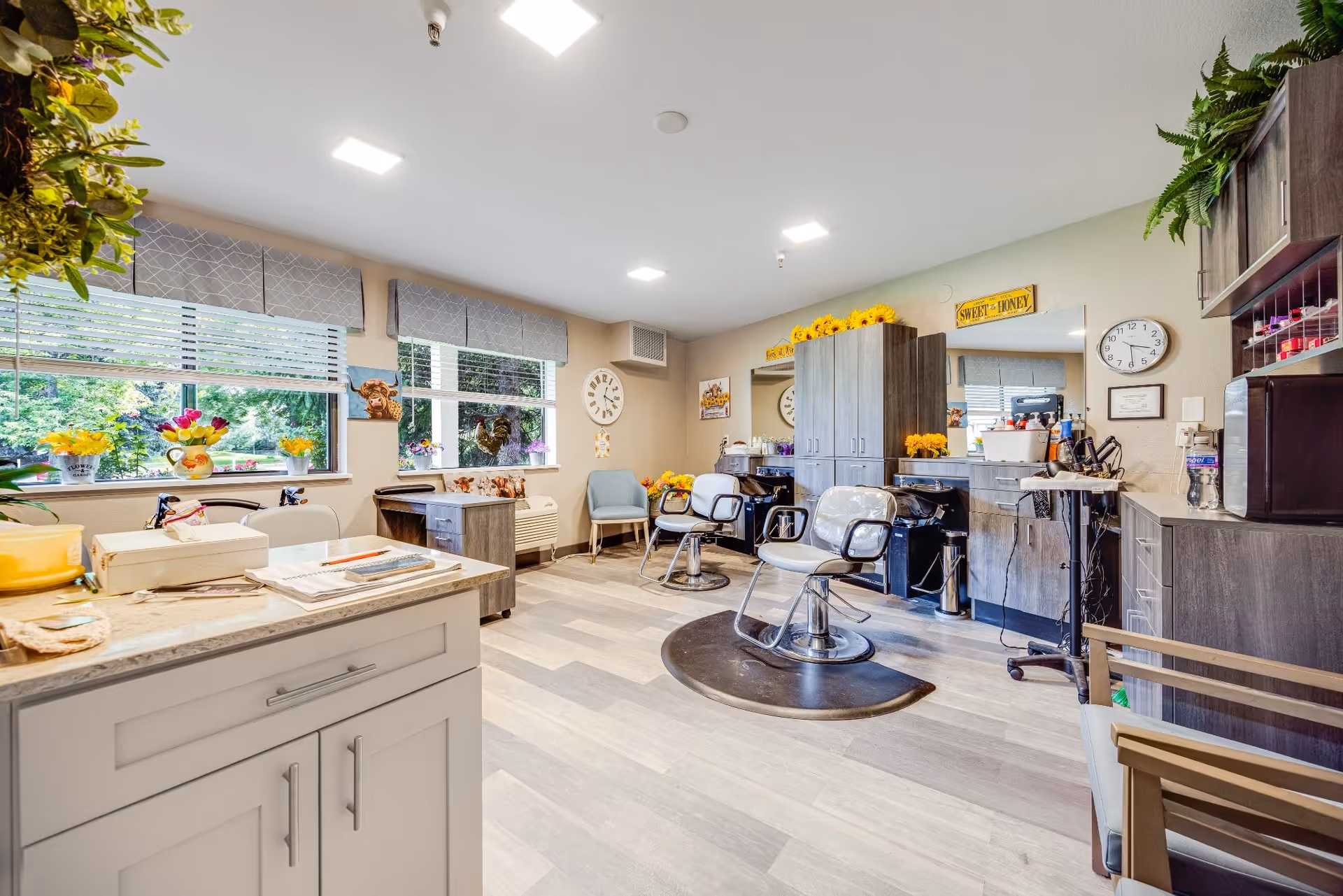A bright and clean salon room with two styling chairs on black mats, large mirrors, and wooden cabinets. The room has large windows with blinds and valances, decorated with flowers and small animal figurines. There are chairs along the walls and various salon equipment and supplies on countertops and shelves. The walls are light-colored with decorative signs and a clock.