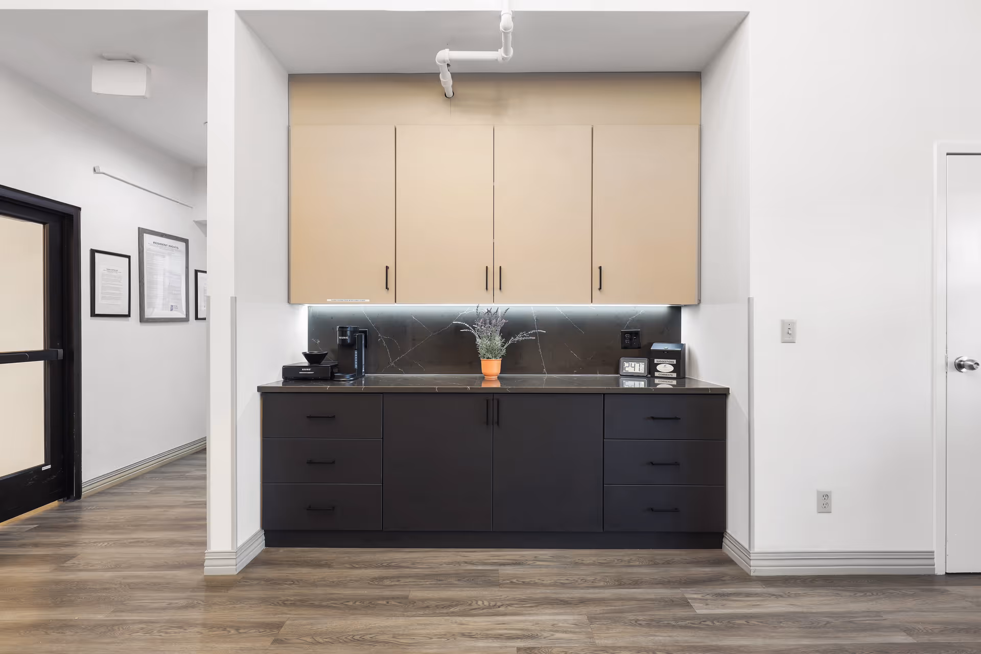 Small kitchenette with beige upper cabinets, dark lower cabinets, a countertop coffee maker and potted plant in a hallway.