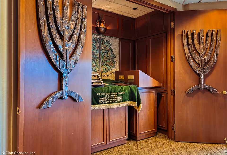 Interior view of a synagogue room with wooden paneling and two large metal menorah decorations mounted on the doors. Inside the room, there is a wooden podium and a table covered with a green cloth embroidered with gold text. A decorative mosaic of a tree is visible on the back wall.