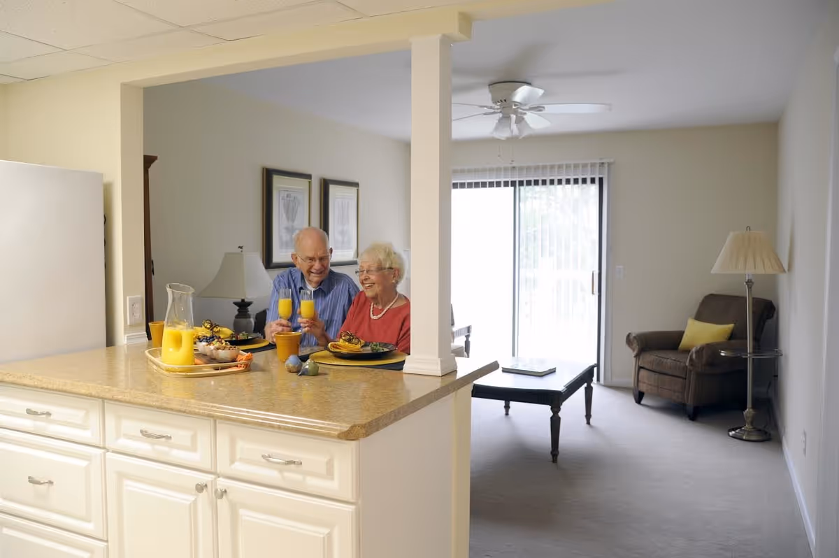 An elderly couple sitting at a kitchen counter enjoying breakfast with orange juice and food. The kitchen counter has a pitcher of orange juice and a tray with snacks. In the background, there is a living room area with a brown armchair, a floor lamp, a coffee table, and a sliding glass door with vertical blinds.