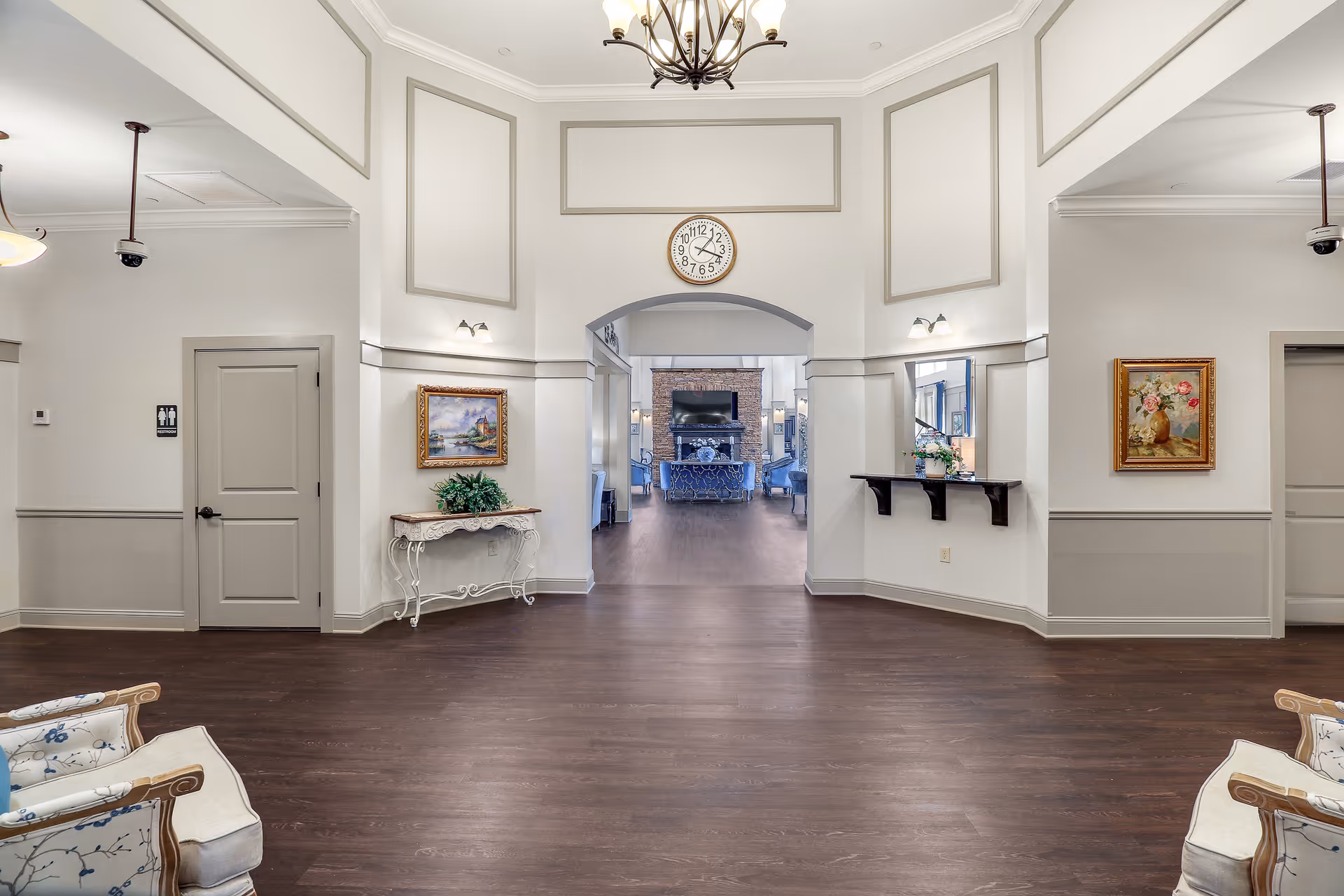 A spacious, well-lit senior living facility interior with high ceilings and a large clock above an arched doorway. The room features light-colored walls with decorative molding, two framed paintings, a small ornate table with a plant, and a shelf with flowers. Two armchairs with floral upholstery are partially visible in the foreground. Beyond the archway, a cozy sitting area with a fireplace and TV is visible.