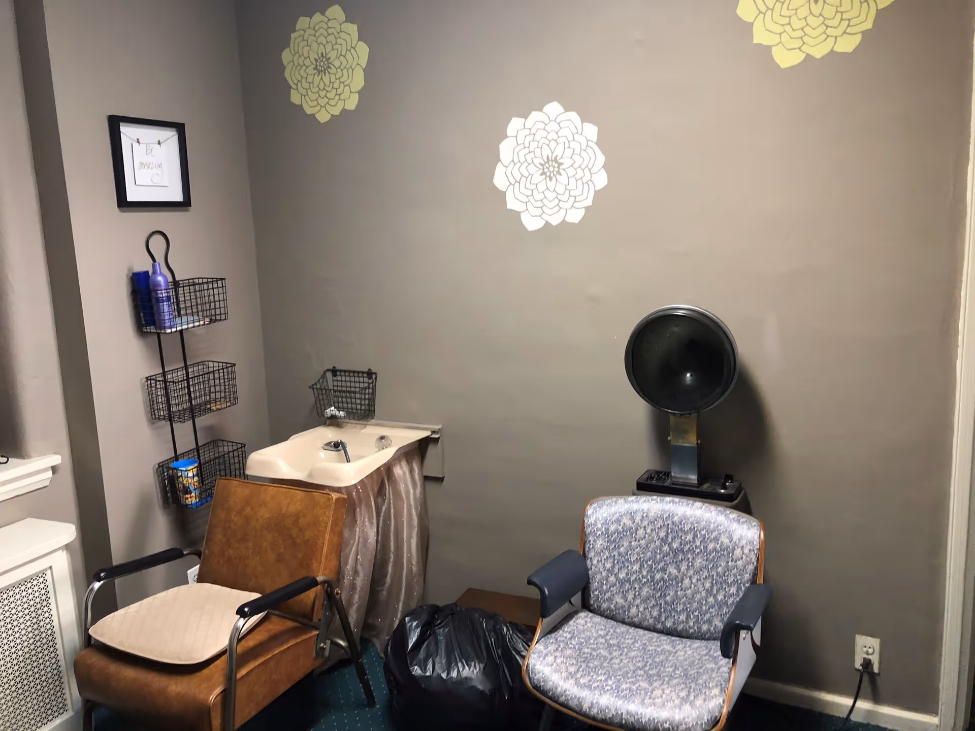 A small room with two chairs, one brown with a cushion and one patterned blue, positioned near a beige sink with a curtain underneath. On the wall above the sink are three decorative flower decals in white and yellow. A black hair dryer hood is mounted on a stand next to the blue chair. A black wire rack with three shelves holding various items is attached to the wall on the left side. The walls are painted gray and the floor is carpeted in green.