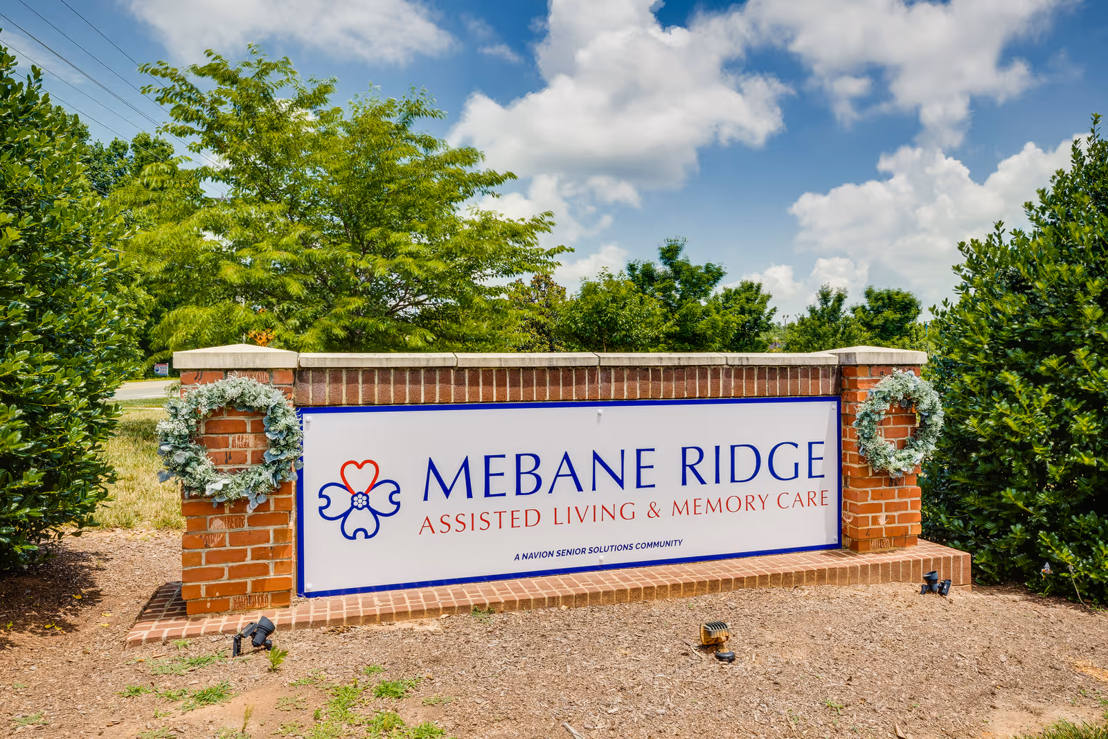 A brick sign for Mebane Ridge Assisted Living & Memory Care, decorated with two wreaths on either side, surrounded by greenery and under a partly cloudy sky.