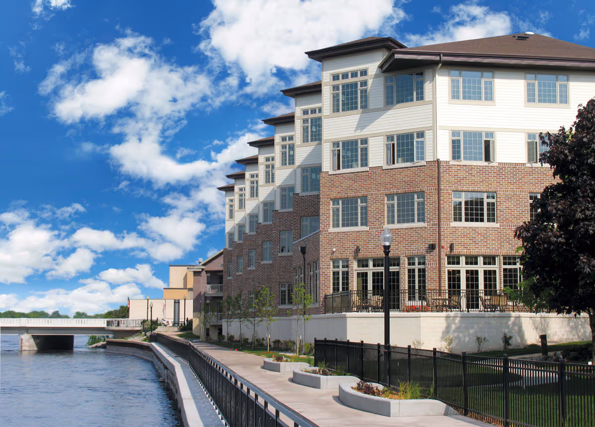 Exterior view of a multi-story senior living facility building with brick and white siding, large windows, and a patio area overlooking a river with a walkway and black metal railing along the water. A bridge is visible in the background under a partly cloudy blue sky.