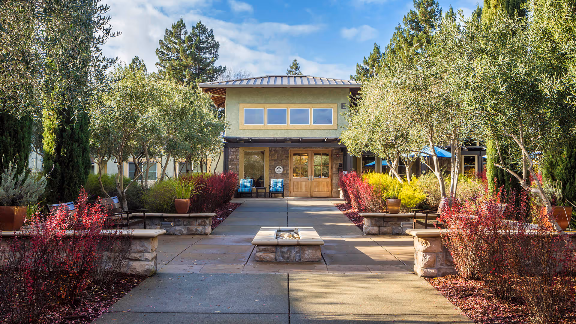 Outdoor courtyard area at Atria Tamalpais Creek featuring a paved walkway lined with stone benches, planters with red and yellow foliage, and small trees leading to a building entrance with double wooden doors and windows above. The sky is partly cloudy and the scene is well-lit with natural daylight.